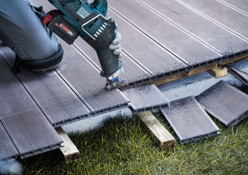 Eine Person mit Sicherheitsausrüstung verwendet ein Elektrowerkzeug, um Terrassendielen im Freien zu schneiden. Anwendungsbild