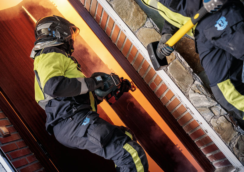 Eine Person mit Sicherheitsausrüstung verwendet ein Elektrowerkzeug, um eine Tür vor einem Feuer zu öffnen. Anwendungsbild